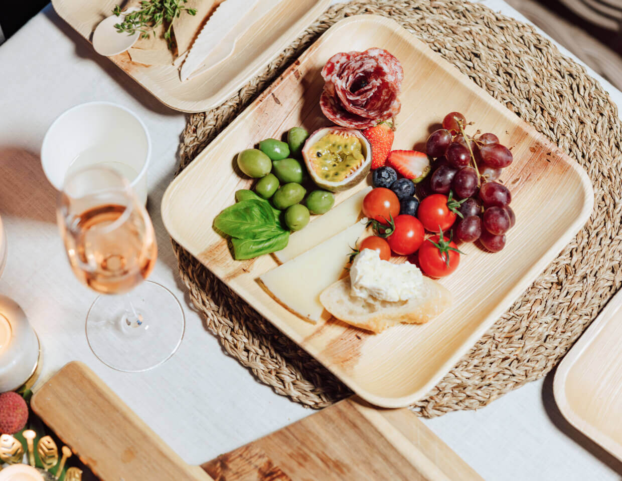 Biodegradable ChicLeaf Palm Leaf Plates & Trays with assorted fruits, cheese, and a glass of rose wine on a white table.