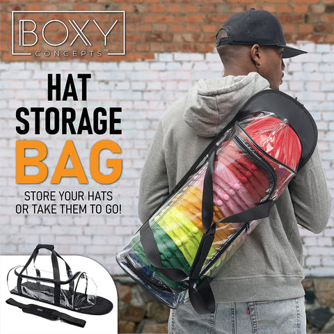 Person carrying a transparent Boxy Concepts' hat storage bag filled with colorful hats against a brick wall.