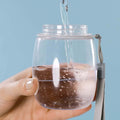 MalsiPree's pet water bottle without it's attached bowl cap head, being filled up with water, held by a hand against a light blue background.